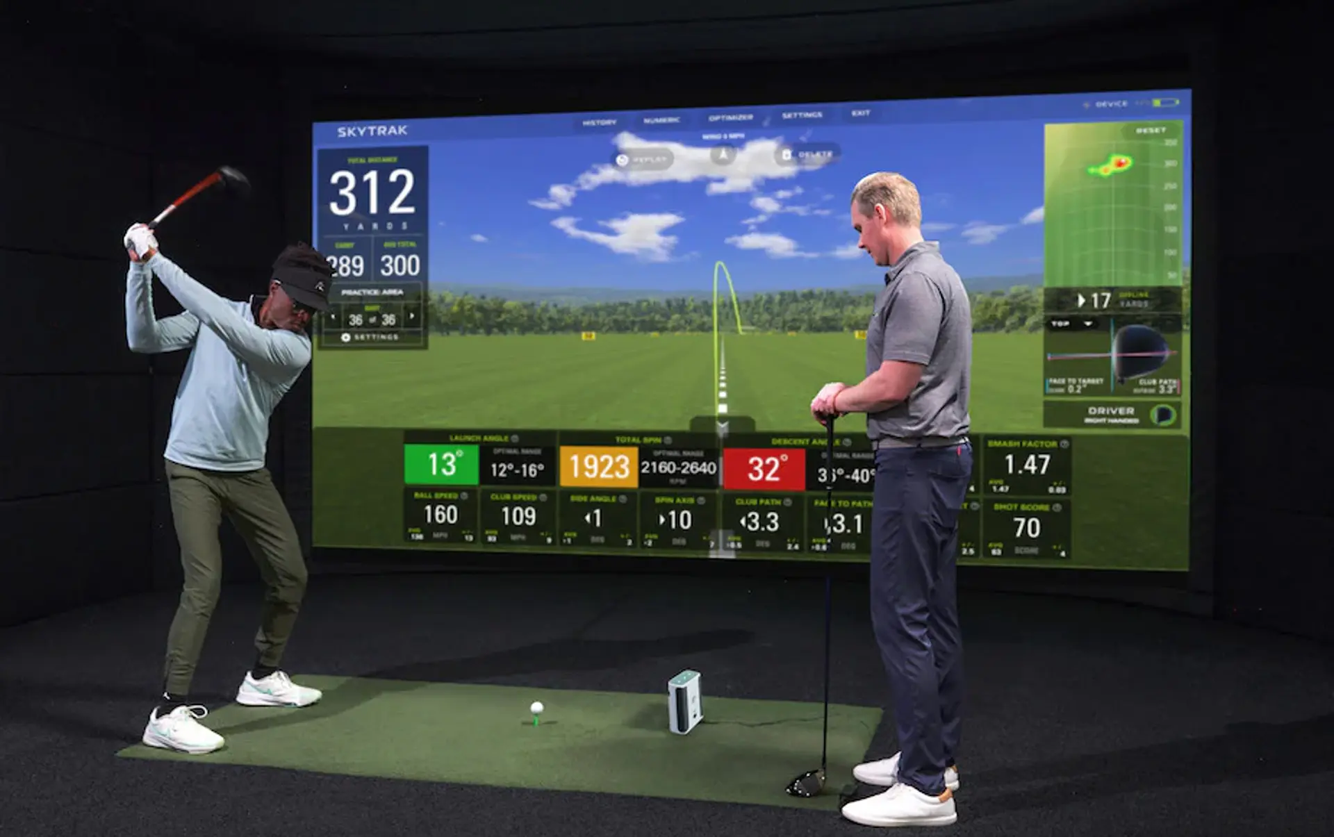 Två personer använder Hitness golfsimulator inomhus, där en golfspelare är på väg att slå ett slag.