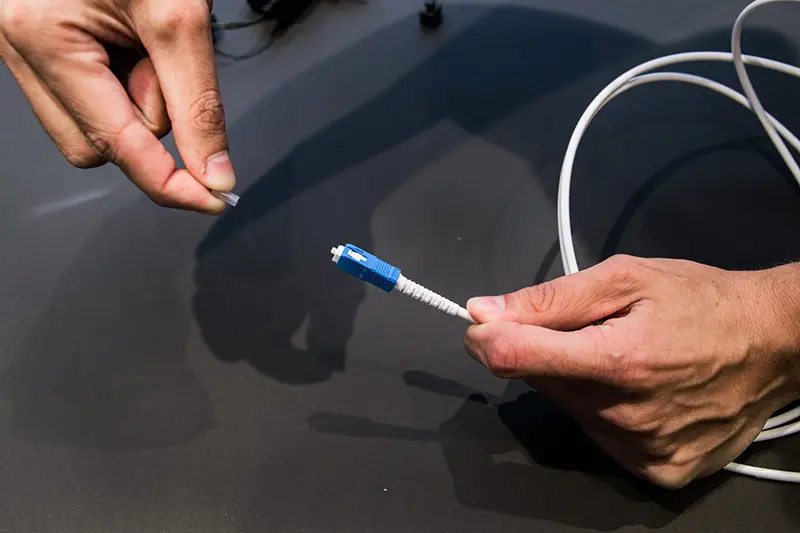 Person som tar bort skyddshatt från blå fiberkontakt på en vit fiberkabel inför anslutning, mot mörk bakgrund.
