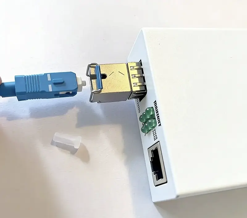 Närbild på blå fiberkabel som ska anslutas till SFP-modul i en vit mediakonverter, med skyddshatt borttagen och LED-lampor samt nätverksuttag synliga på framsidan.