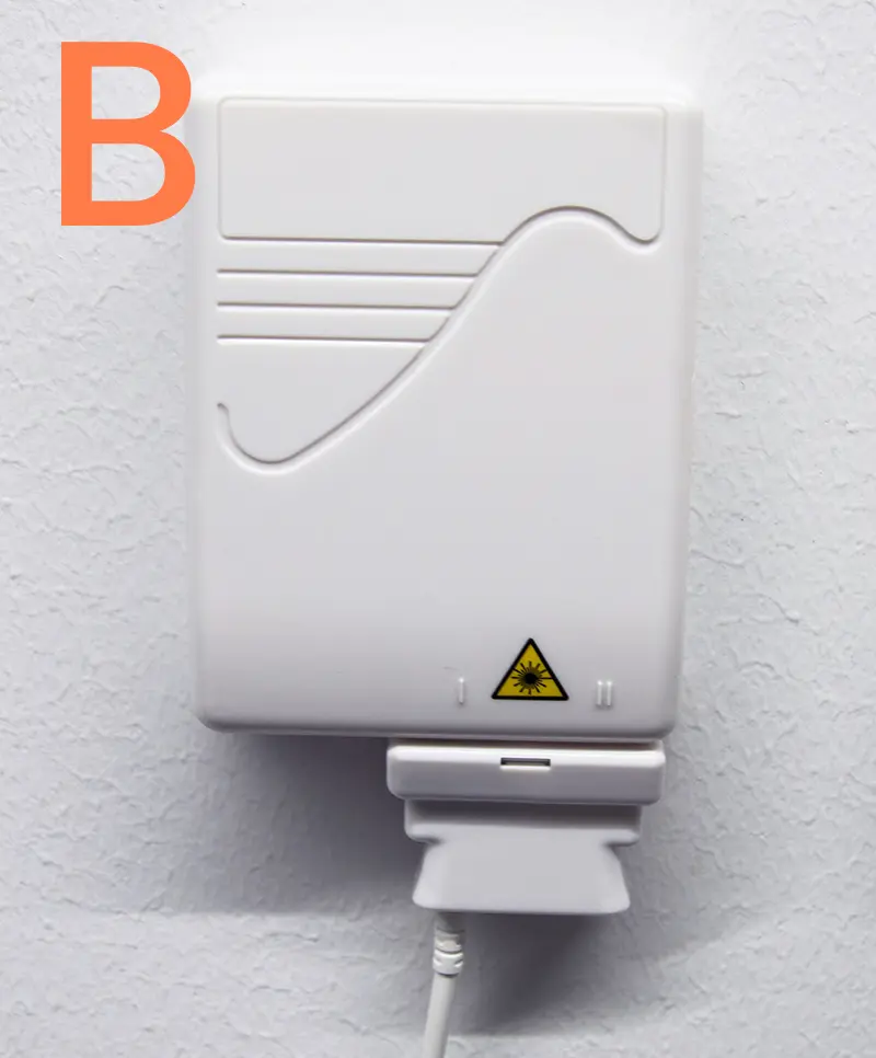 Fiberuttag modell B monterat på vit vägg, med vågformad front, lasersymbol och ansluten fiberkabel nedtill.